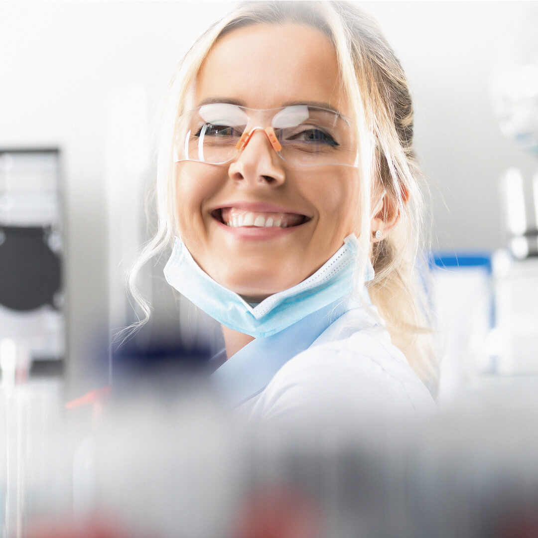 Das Bild zeigt eine junge Chemikantin, die in die Kamera lächelt. Sie trägt einen Mundschutz, eine Schutzbrille und einen weißen Kittel