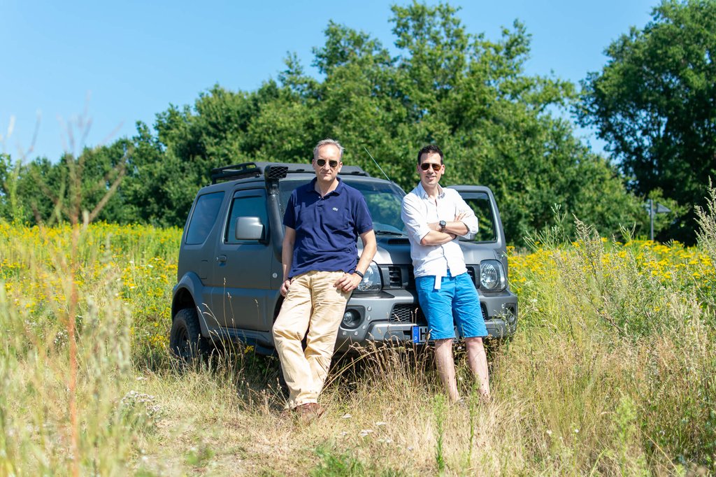 Moritz von Grotthuss und Sascha Klement lehnen an ihrem Auto, das auf einer Wiese steht. Beide tragen eine Sonnenbrille und blicken in die Kamera.