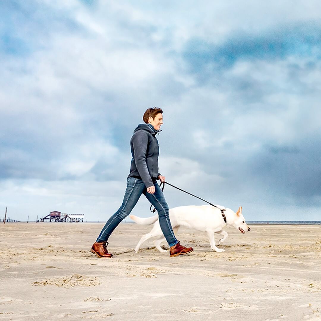 Eine Frau läuft mit ihrem Hund an der Leine entlang eines Strands, der Himmel ist blau und mit grauen Wollen durchzogen.