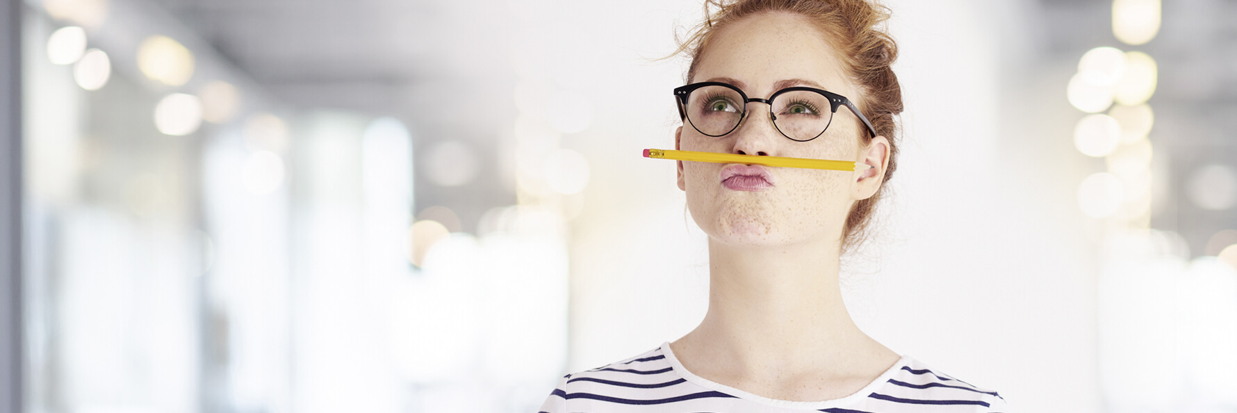 Eine junge Frau hält zwischen Nase und Oberlippe einen gelben Bleistift. Sie sieht dabei nach oben und macht einen grübelnden Gesichtsausdruck.